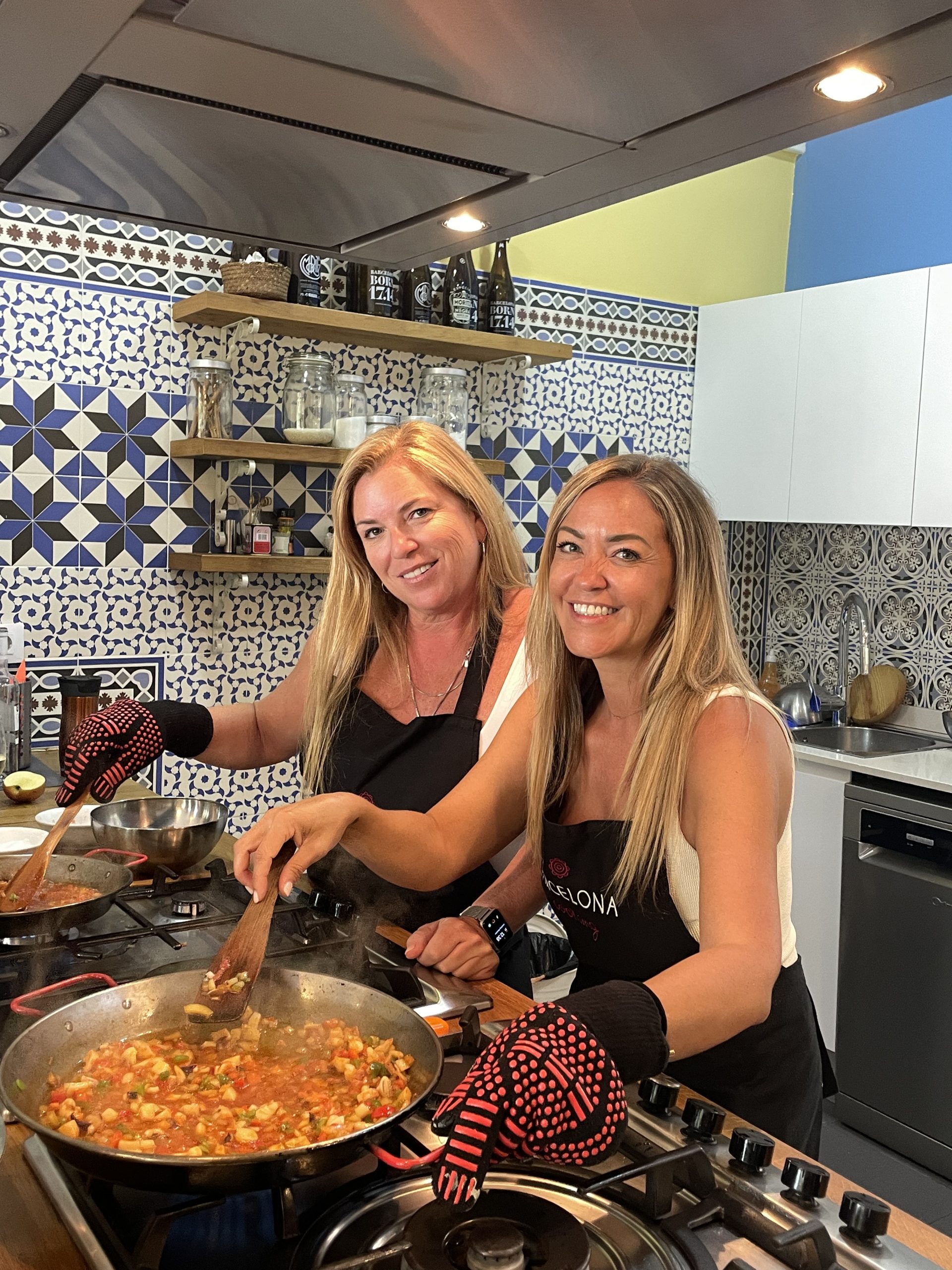 Two women preparing a meal together in a modern kitchen, smiling while cooking at a stovetop with a patterned tile backsplash